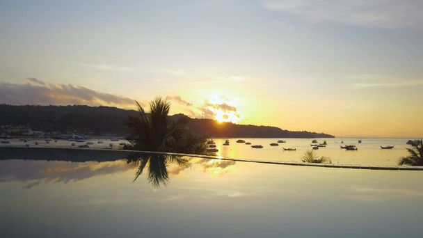 LENS FLARE: Henkeäsalpaava kultainen auringonlasku loistaa alas viileä tyhjä ääretön uima-allas näkymät hiekkaranta täynnä palmuja. Viehättävä trooppinen kesäilta rauhallisella lahdella Phi Phi saarilla
. - Valokuva, kuva