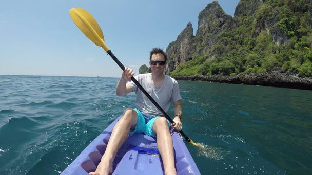CLOSE UP PORTRAIT: Young male having fun paddling his kayak in picturesque Phi Phi Islands. Caucasian man looks at stunning limestone karst islands and turquoise ocean water while kayaking in Thailand - Photo, Image