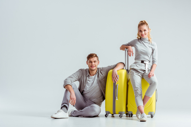 couple sitting near yellow suitcases and looking at camera on grey background, travel concept - Фото, зображення