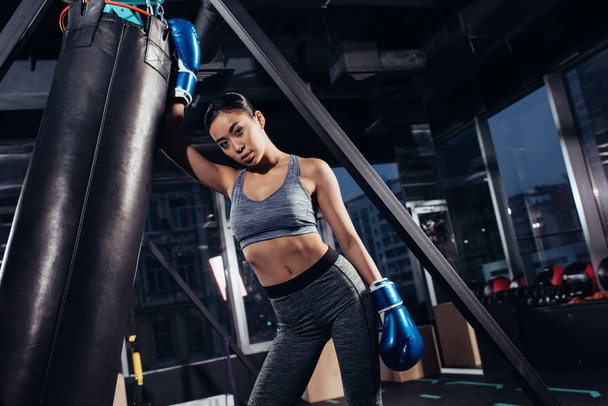 sexy asian girl in boxing gloves standing near punching bag at gym - Foto, Imagen