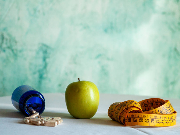 Яблуко, міра стрічки і синя ємність з таблетками рослинного волокна на столі
 - Фото, зображення