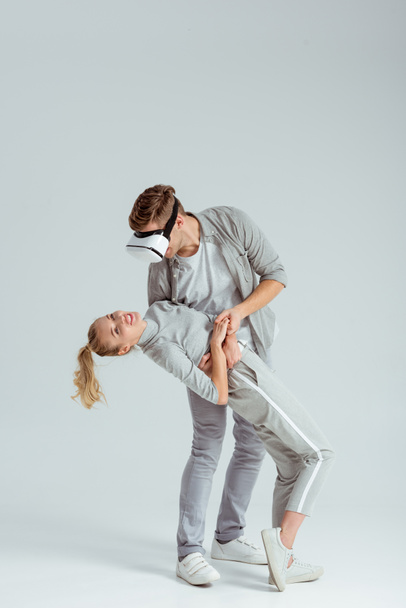 hombre sosteniendo mujer en abrazo mientras tiene experiencia de realidad virtual sobre fondo gris
 - Foto, Imagen