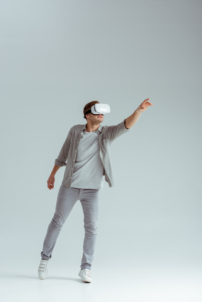 man in grey clothing gesturing while wearing virtual reality headset on grey background  - Foto, Imagem