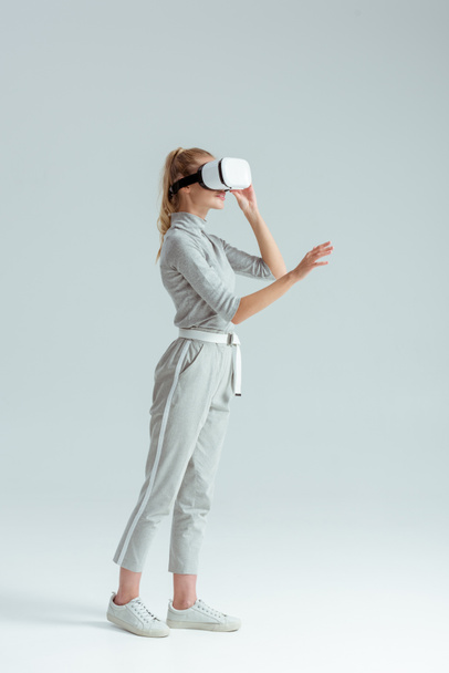 girl in grey clothing and vr headset gesturing while having virtual reality experience on grey background - Photo, Image