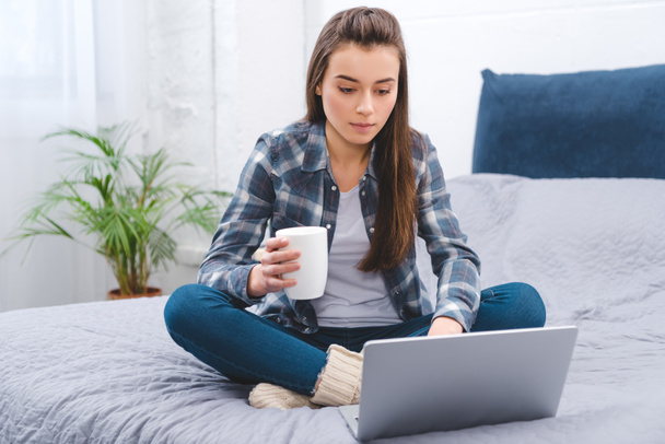 beautiful girl holding mug with hot beverage and using laptop on bed  - Photo, Image