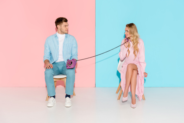 smiling woman having conversation while man holding purple vintage telephone on pink and blue background - Φωτογραφία, εικόνα