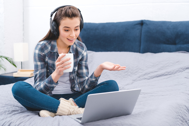smiling young woman in headphones holding cup and chatting with laptop in bedroom  - Фото, зображення