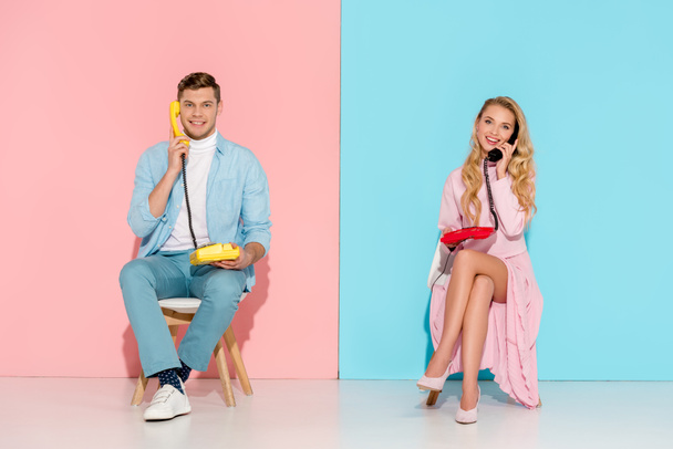 smiling couple sitting and having conversation on vintage telephones with pink and blue background - Foto, afbeelding