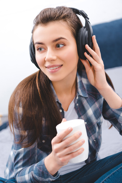 attractive smiling girl in headphones holding cup and looking away at home - Foto, Imagen