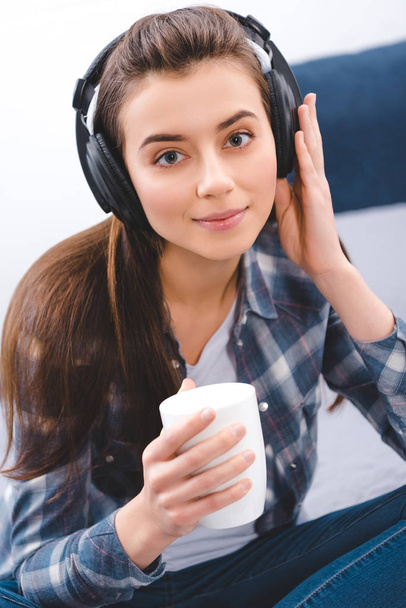 beautiful young woman in headphones holding cup and smiling at camera - Φωτογραφία, εικόνα