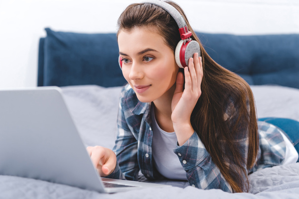 beautiful smiling young woman in headphones using laptop while lying on bed  - Φωτογραφία, εικόνα