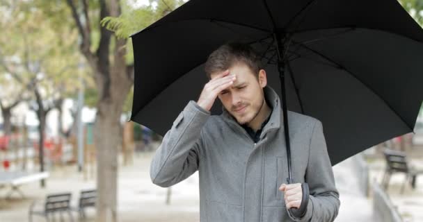 Angry man suffering head ache walking towards camera holding an umbrella under the rain in a park - Filmagem, Vídeo