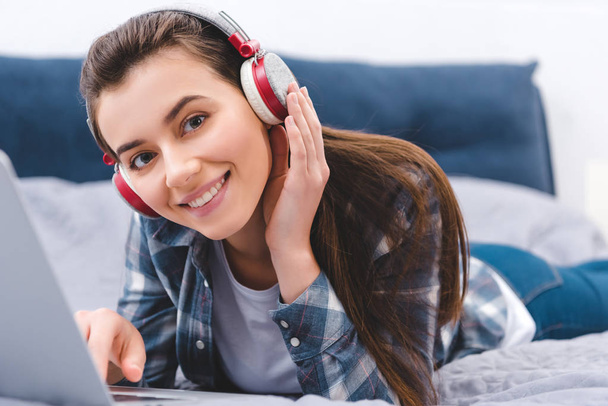 attractive girl in headphones using laptop on bed and smiling at camera   - Fotoğraf, Görsel