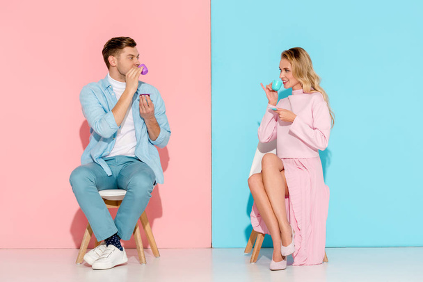 mooie paar zitten op stoelen, kijken naar elkaar en koffie drinken op roze en blauwe achtergrond - Foto, afbeelding