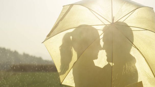 Silueta Close Up odlesk objektivu: Muž a žena v lásce schovávat za deštník, jak se chystá políbit zlaté venkově. Veselý mladý pár tančí v dešti se zastaví a hledí na sebe. - Fotografie, Obrázek