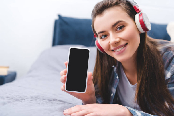 houkutteleva tyttö kuulokkeet tilalla älypuhelin tyhjä näyttö ja hymyilee kameralle, kun makaa sängyssä
  - Valokuva, kuva