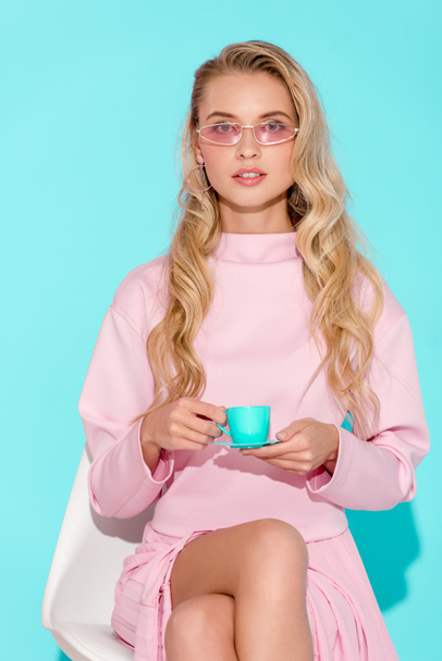 beautiful woman in pink outfit and sunglasses holding toy coffee cup and looking at camera on turquoise background - Photo, Image