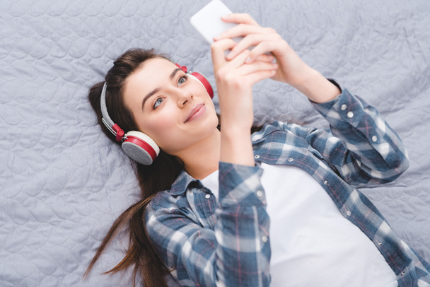 beautiful smiling girl in headphones lying on bed and using smartphone - Fotó, kép
