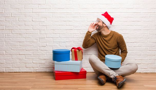 Junger Mann sitzt mit Geschenken bei der Weihnachtsfeier und flüstert Klatsch-Unterton - Foto, Bild