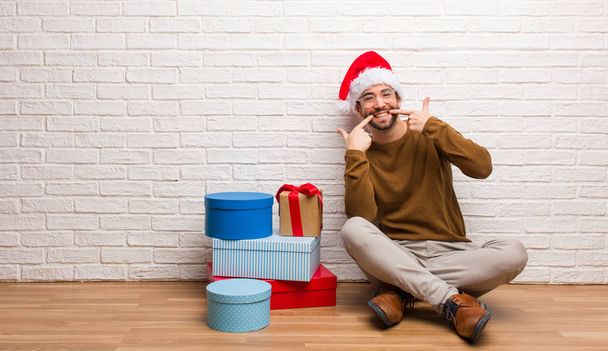Молодий чоловік сидить з подарунками, святкуючи різдвяні посмішки, вказуючи рот
 - Фото, зображення