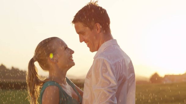 LENS FLARE, CLOSE UP: Easygoing Caucasian couple laughs while they dance alone in stunning nature on a rainy summer evening. Cinematic shot of young boyfriend and girlfriend enjoying the spring rain. - 写真・画像