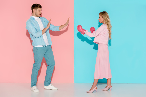 man gesturing with hands near aggressive woman in pink boxing gloves on pink and blue background - Photo, Image