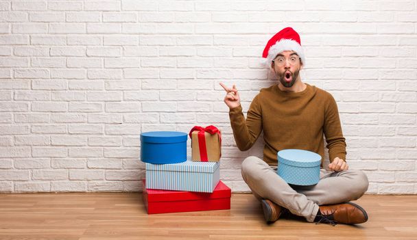 Young man sitting with gifts celebrating christmas pointing to the side - Фото, изображение