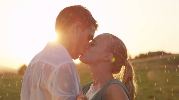 CLOSE UP, LENS FLARE: Beautiful blonde woman and handsome man on a lovely date outdoors kiss in the summer rain. Carefree young couple gets wet as they twirl and kiss in a refreshing summer shower - Photo, Image