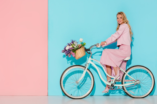 smiling woman in pink clothing riding bicycle with flower basket on pink and blue background - Фото, зображення