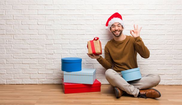 Young man sitting with gifts celebrating christmas cheerful and confident doing ok gesture - Foto, afbeelding