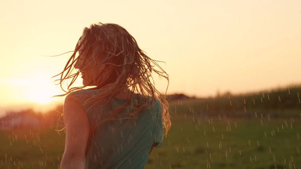 Close Up, αναλαμπή φακού: Ξέγνοιαστες ξανθιά γυναίκα χορεύει σε εξωτερικούς χώρους στη βροχή φρέσκα άνοιξη στο ηλιοβασίλεμα. Πανέμορφο κορίτσι περιστροφές παιχνιδιάρικα στην όμορφη ύπαιθρο σε ένα μαγευτικό πορτοκαλί ανάβει καλοκαιρινό βράδυ. - Φωτογραφία, εικόνα