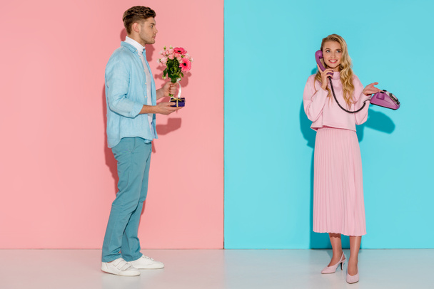 чоловік тримає квітковий букет і подарункову коробку, коли усміхнена жінка розмовляє на старовинному телефоні з рожевим і синім фоном
 - Фото, зображення