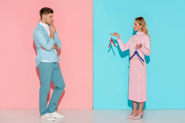 thoughtful man looking at woman and choosing empty clothes hangers on pink and blue background - Foto, Imagen