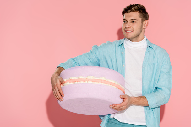 handsome man in blue clothing holding big macaroon model on pink background - Photo, Image