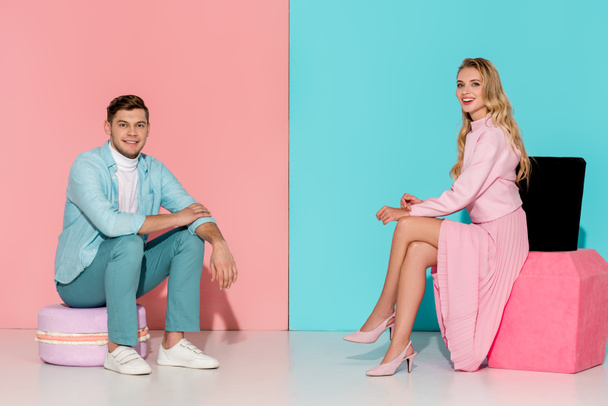 couple sitting on big macaroon and nail polish models while looking at camera on pink and blue background - Foto, afbeelding