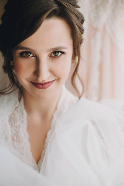 Beautiful stylish brunette bride posing in silk robe under veil in the morning. Happy woman model with perfect makeup and hairstyle, getting ready for wedding day - Foto, afbeelding