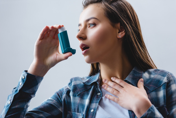 young woman using asthma inhaler and looking away isolated on grey - Photo, Image