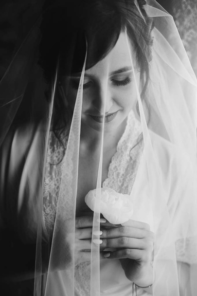 Beautiful stylish brunette bride posing in silk robe under veil in the morning. Sensual portrait of happy woman model with peony flower in hands getting ready for wedding day - Photo, image