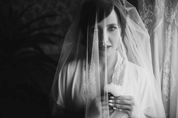 Mooie stijlvolle brunette bruid poseren in zijde gewaad onder sluier in de ochtend. Sensuele portret van gelukkige vrouw model met corsages in handen klaar voor wedding day - Foto, afbeelding