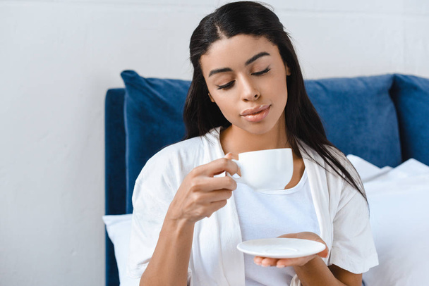 красивая смешанная раса девушка в белом халате пьет кофе в постели утром
 - Фото, изображение