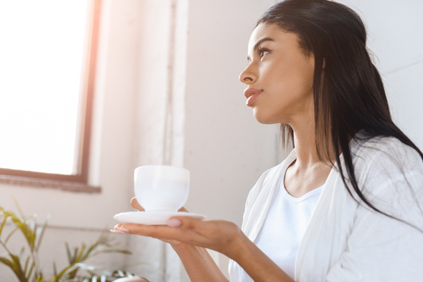 gyönyörű kilátással vegyes verseny lány gazdaság csésze kávé reggeli napfény hálószoba fehér köntöst - Fotó, kép