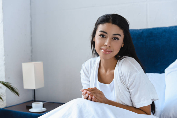 beautiful mixed race girl in white robe sitting on bed and looking at camera in morning - Photo, Image