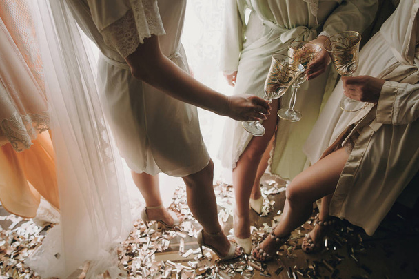 Noiva com damas de honra em roupões de seda brindar com taças de champanhe e mostrando pernas sexy, de pé em ouro e prata confetti, festa da manhã boudoir nupcial. Chuveiro de galinhas
. - Foto, Imagem