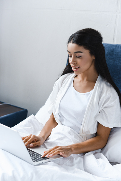 smiling attractive mixed race girl in white robe using laptop in morning in bed - Photo, Image