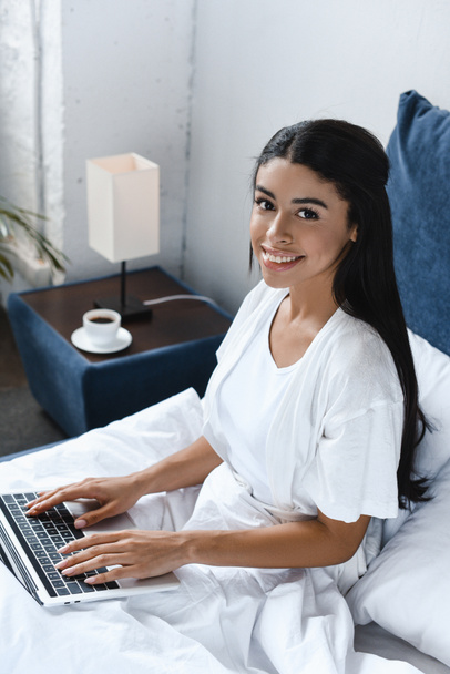 high angle view of smiling beautiful mixed race girl in white robe using laptop in morning in bedroom and looking at camera - Фото, зображення