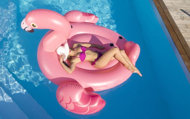 Antenne: junge Frau mit hinreißendem Körper zieht ihren stylischen weißen Hut über die Augen, während sie sich auf einem großen Flamingo-Floatie sonnt. attraktive Touristin im rosa Bikini sonnt sich auf aufblasbarem Spielzeug. - Foto, Bild