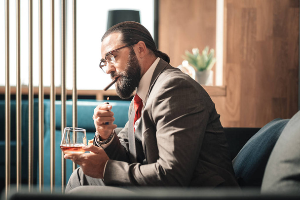 Bearded dark-haired man lighting his cigar while drinking whisky - Photo, Image