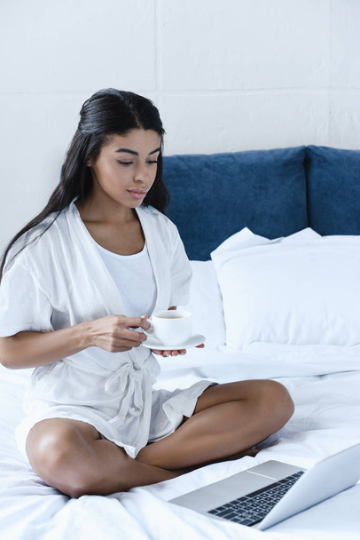 beautiful mixed race girl in white robe holding cup of coffee and looking at laptop in morning in bedroom - Photo, Image