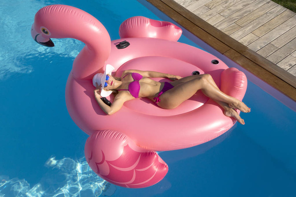 AERIAL: Hermosa chica caucásica sonríe mientras toma el sol en un gran flamenco inflable balanceándose alrededor de la impresionante piscina privada. Alegre turista femenina en bikini rosa se relaja en un flotador de gran tamaño
. - Foto, imagen