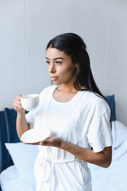 mooi gemengd ras meisje in een witte robe houden van kopje koffie in de ochtend in de slaapkamer en op zoek weg - Foto, afbeelding